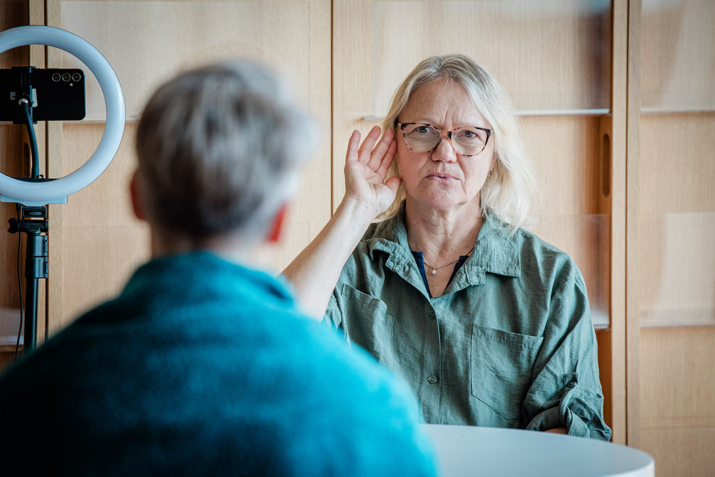 Vaaleahiuksinen ja silmälasipäinen nainen viittoo "kuuleminen". Naista vastapäätä on selin lyhyttukkainen henkilö ja kännykkäteline.